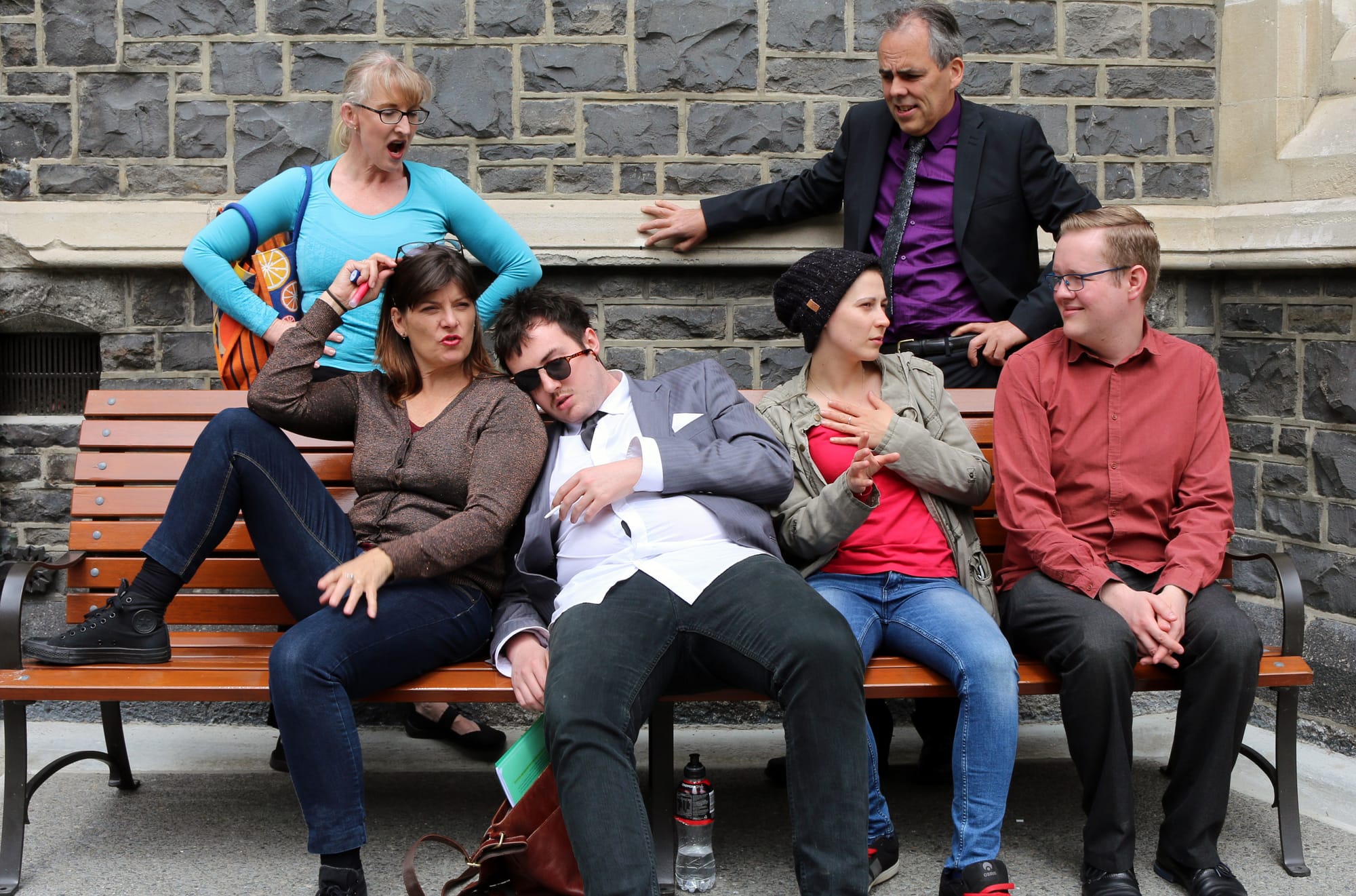 A group of eccentric strangers gather outdoors at a bench.