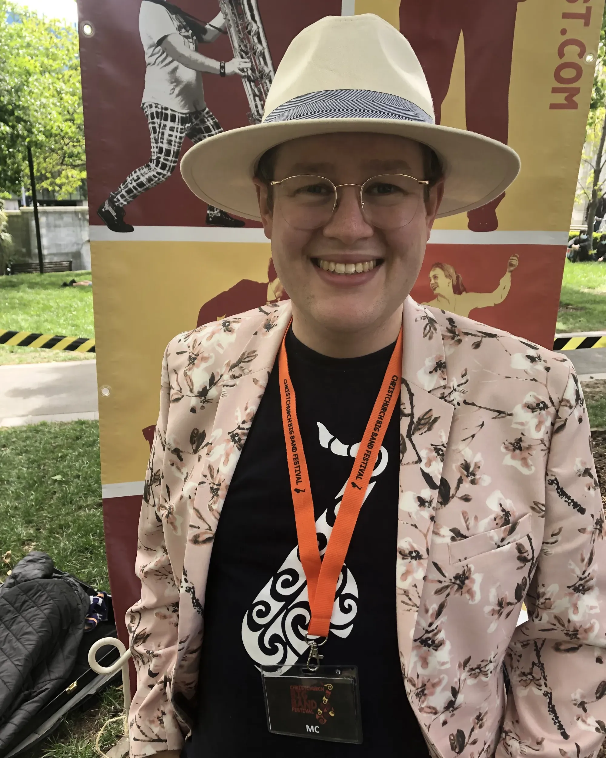 Mark wearing an MC lanyard, a t-shirt with a koru-styled saxphone, a white brimmed hat, and a pale floral suit, standing outside in front of colourful Big Band Festival signage.