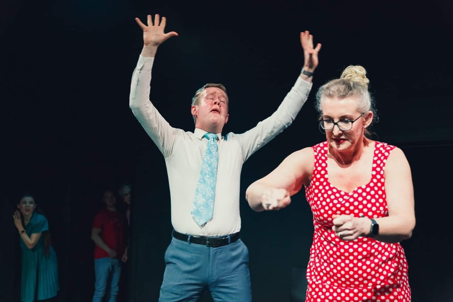 Mark raises his hands in passionate song wearing dress pants, white shirt, and a light blue tie, while a lady in a polkadot dress mimes makes a sandwich.