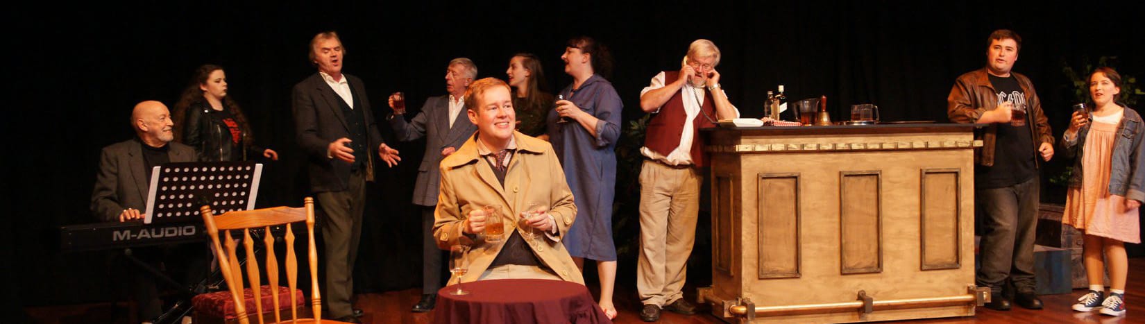 Guy Jones sits at a table holding two drinks uneasily while the townspeople gather at the bar behind him.