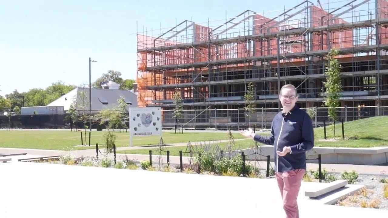 Mark stands on brightly sunlit concrete in front of garden, grass, and a townhouse building site with scaffolding.