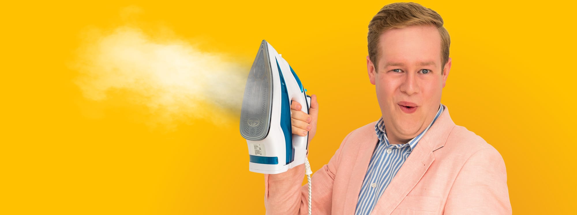 Mark wielding an iron salaciously. He wears a pastel pink suit and a blazer with white and blue stripes. There is a bright yellow background.