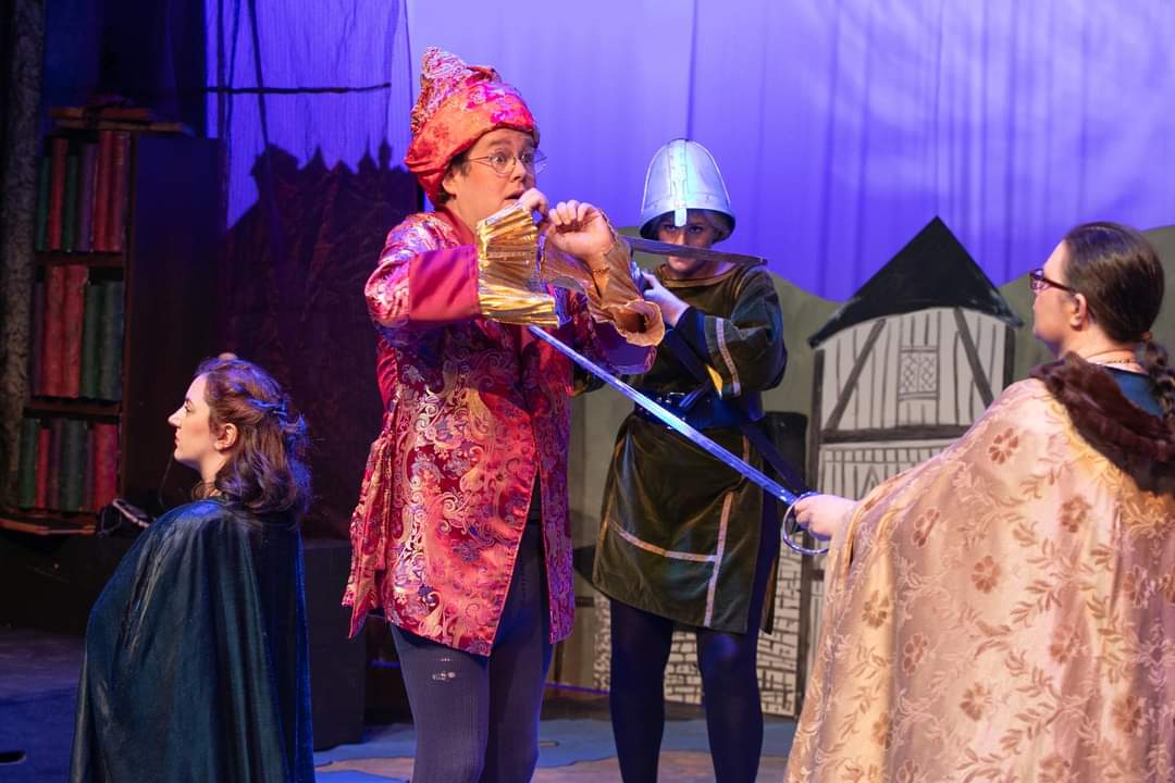 Cutwell the wizard, dressed in tights and a shiny red-gold hat and coat, holds his hands up in fear as a sword is pointed at his chest.