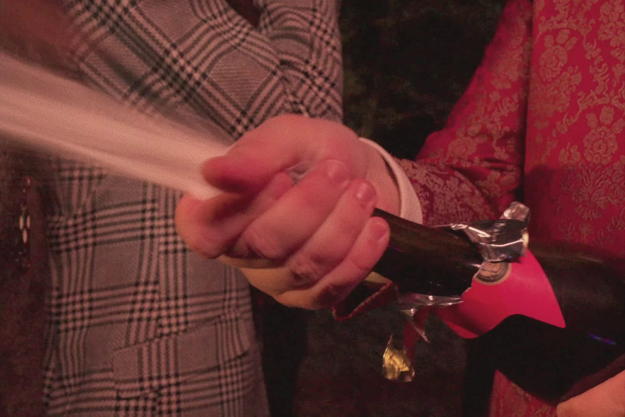 Close-up of of a jet of champagne shooting from a bottle held by a man in a glorious red suit with golden embroidery. In the background is someone wearing a light chequered suit.