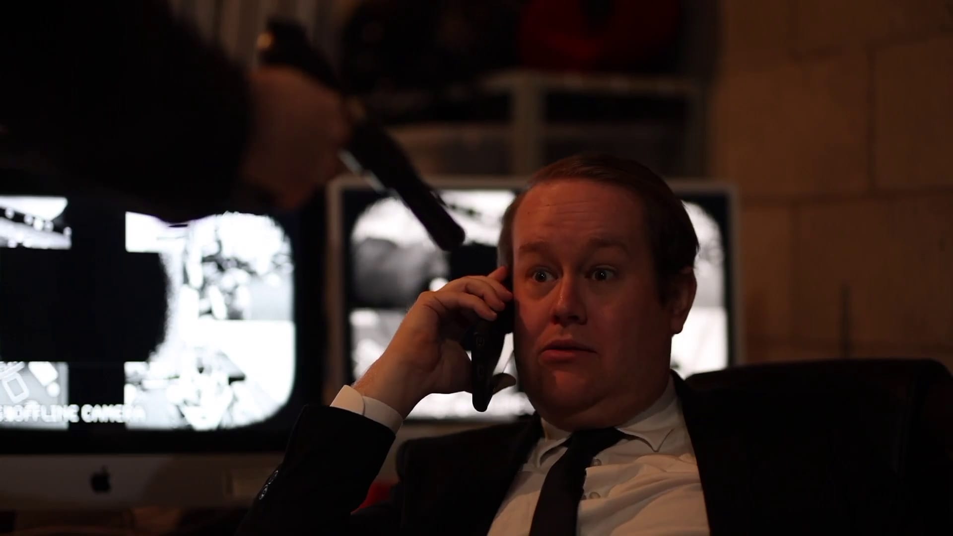 Mark is on the phone wearing a black suit and tie with CCTV screens in the background. He looks very disturbed to have suddenly discovered that there's a gun pointing at him.