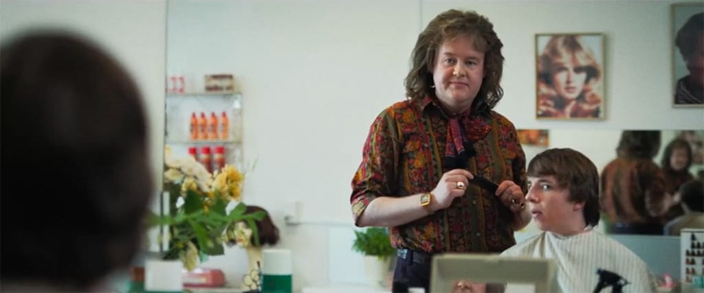 A male 1970s hairdresser and a teenage boy looking in the mirror. The hairdresser has long voluminous hair and is wearing a colourful shirt and tied scarf and an ostentatious ring and digital watch. He is holding a comb. The boy is mid-haircut. He has longish hair and is unsure of the haircut. In the background are hair products, an image of a lady's hairstyle, flowers, and a reflection of a mirror showing a reflection of the reflection.