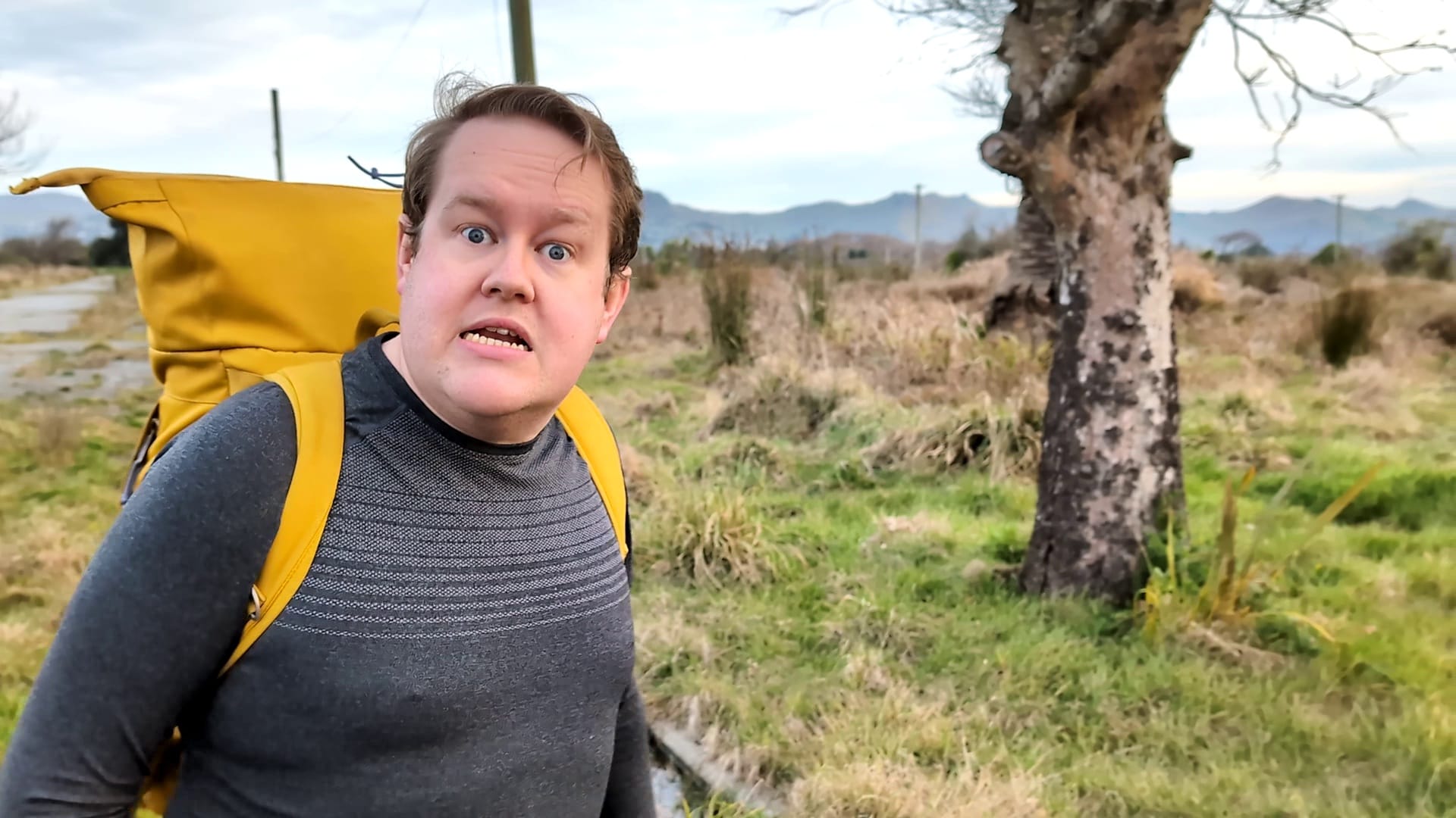 Mark Darbyshire looks at the camera very irately, his jaw protuding, with unkempt grass, overgrown road, and leafless trees of the Christchurch Red Zone behind him. He his wearing a tall bright-yellow backpack.