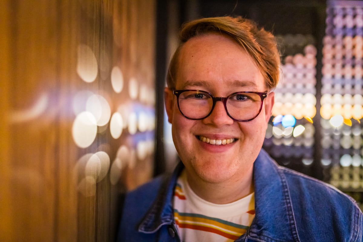 Photo with pretty circles of light. Mark is smiling and wearing glasses, a stripey t-shirt, and a denim jacket.