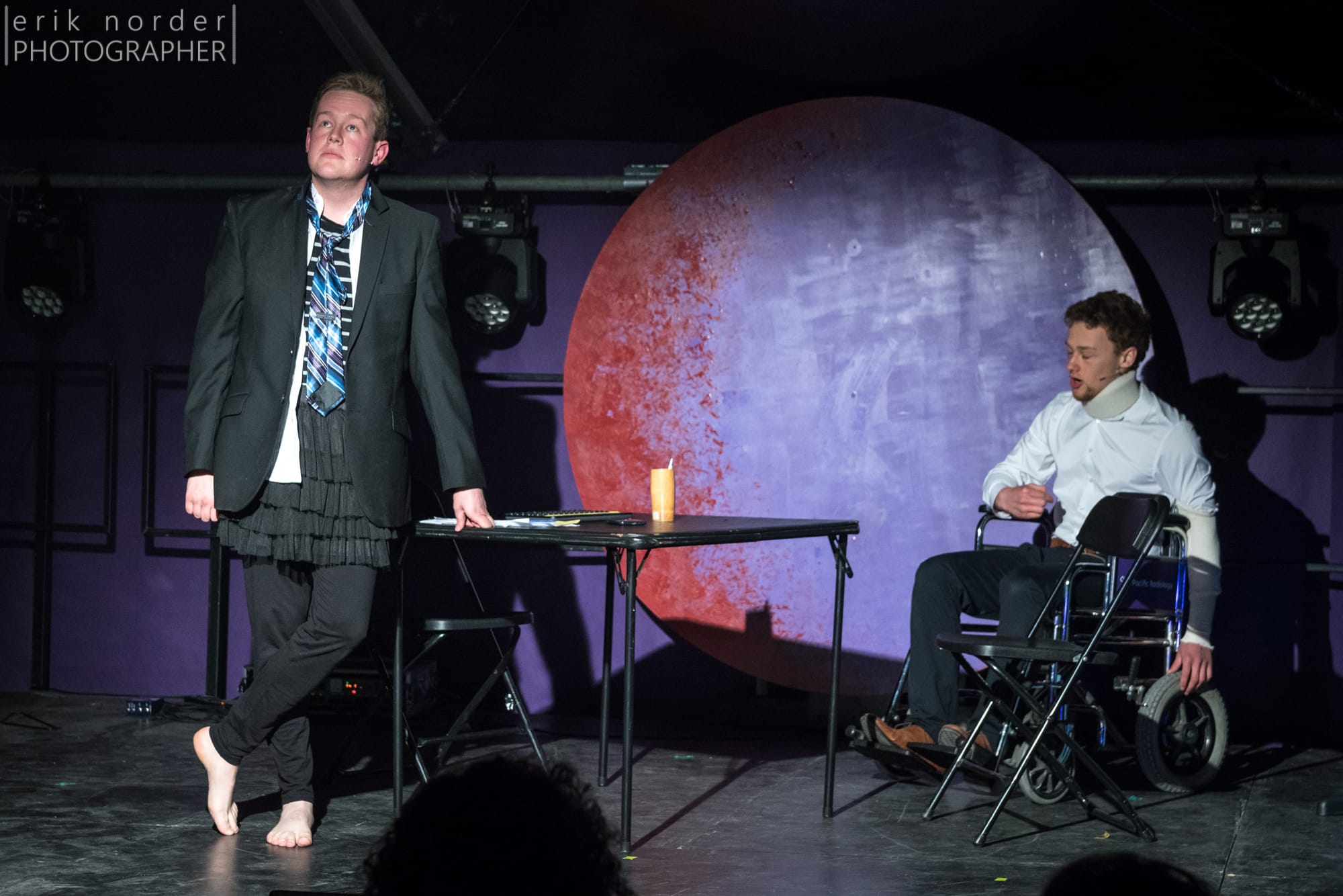 Mark wearing a black blazer, loose striped blue tie, white shirt, black striped skivvy, black tutu, black tights, and bare feet, gazing into the distance. To his right, a man in a wheelchair wearing office attire and a neck brace. By Erik Norder Photographer.