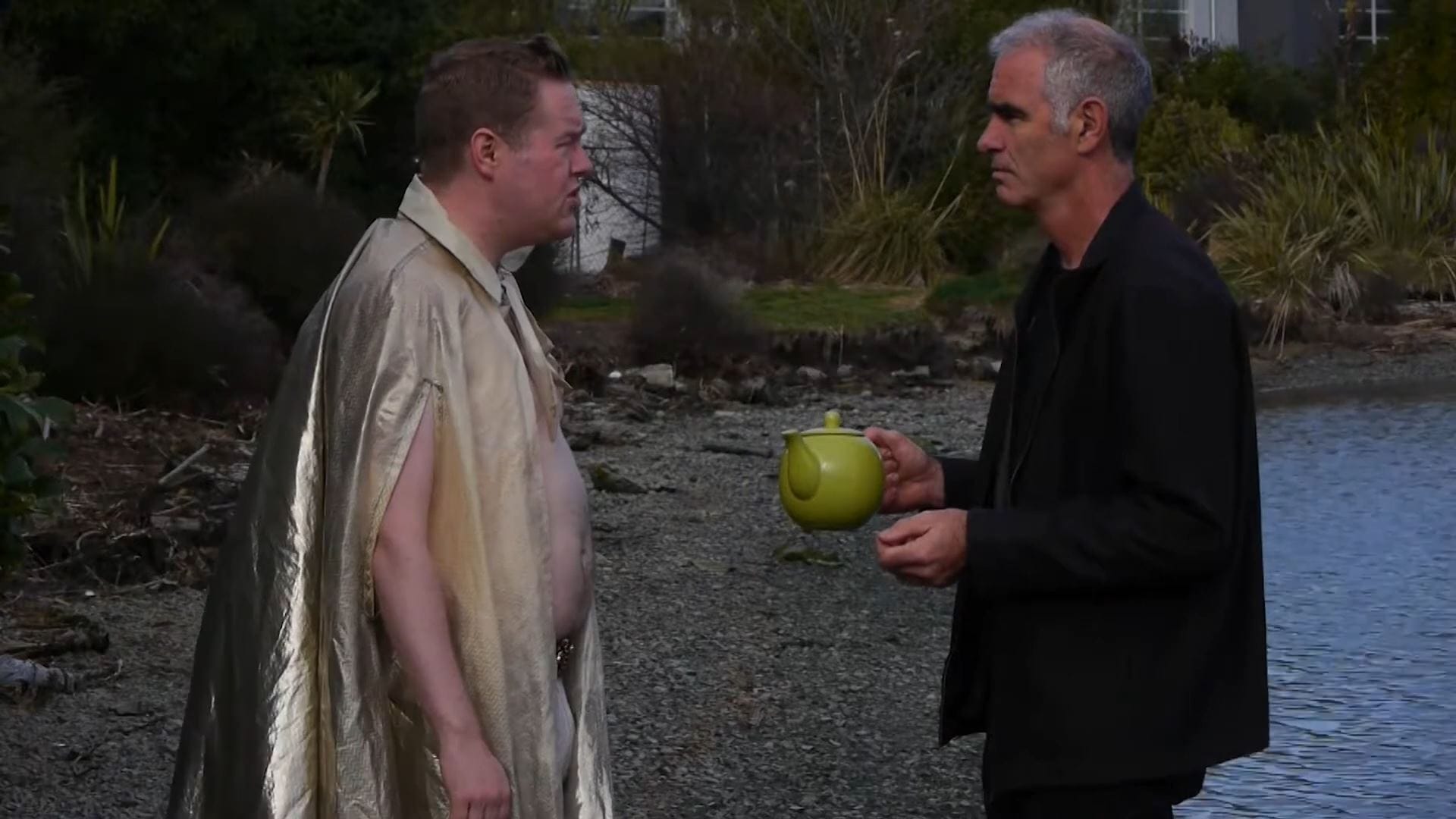 A chubby, tackily dressed genie has just appeared in front of a man holding a teapot on a pebbled shore.