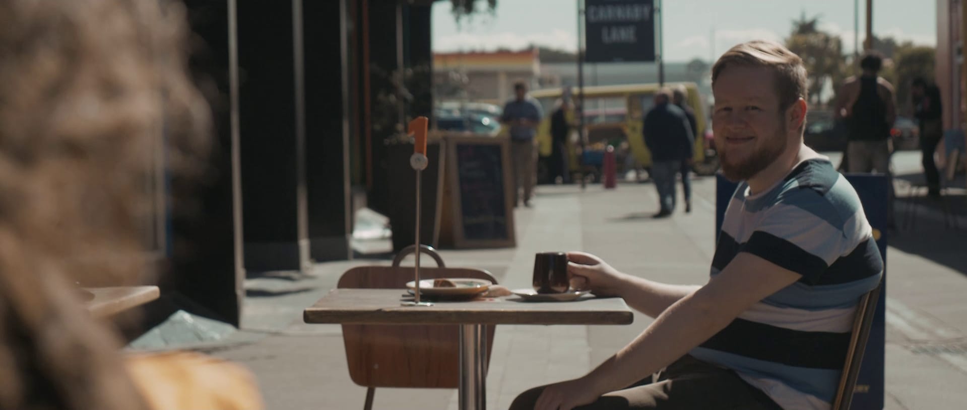 Mark Darbyshire sits with his coffee at an outdoor cafe table and smiles at a stranger just out of shot