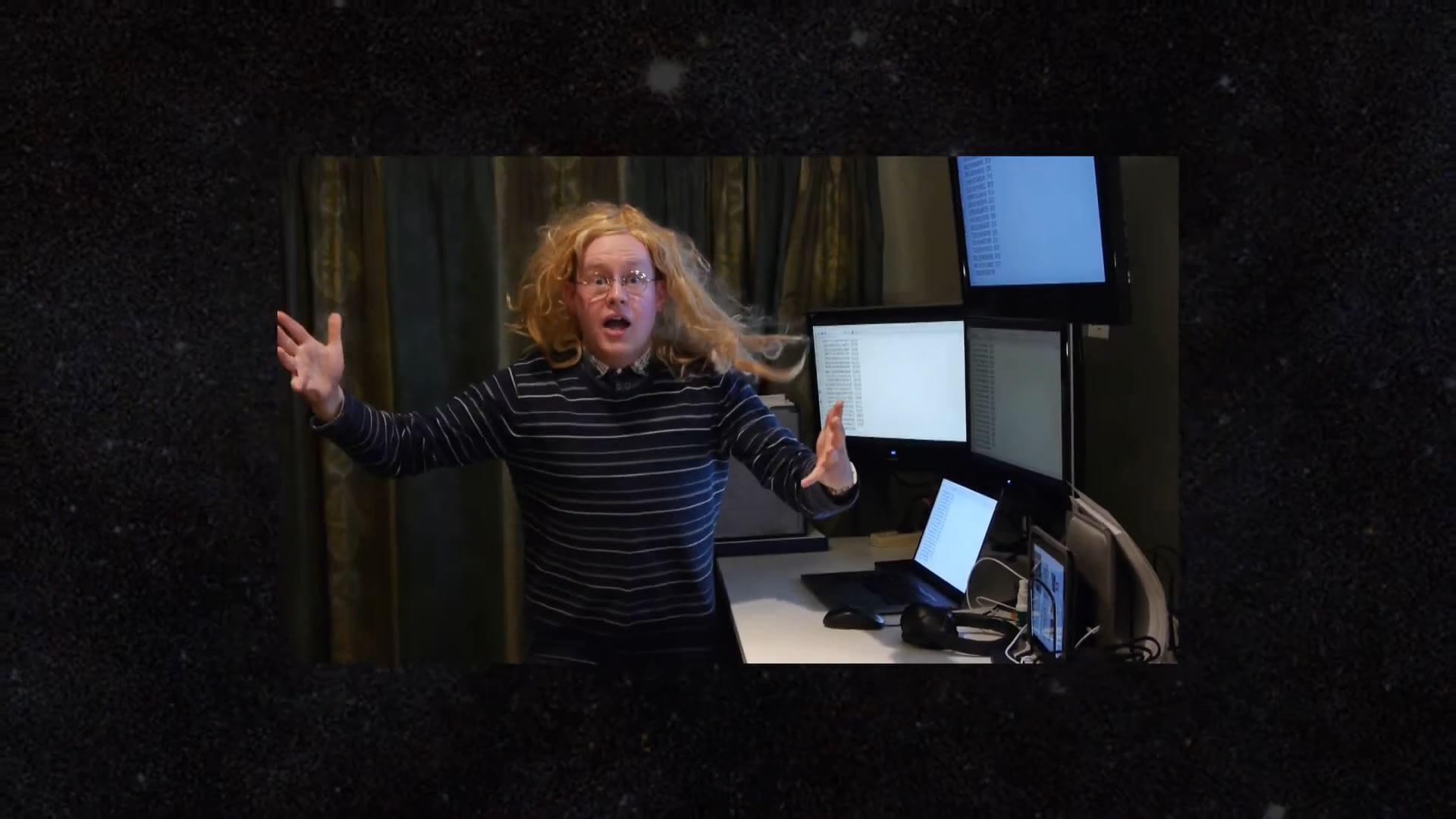 A shot of an excited man with long hair and arms outstretched beside four computer screens is blown away