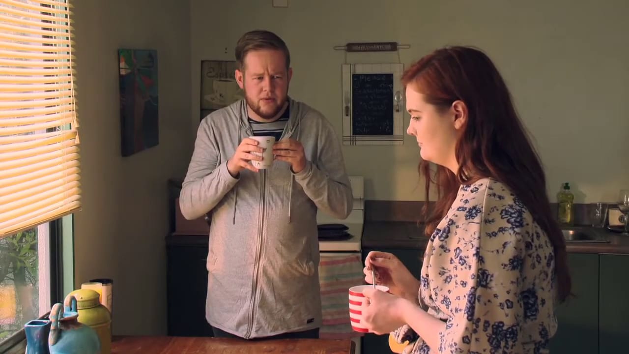 A man and a woman have hot drinks in a kitchen looking concerned.