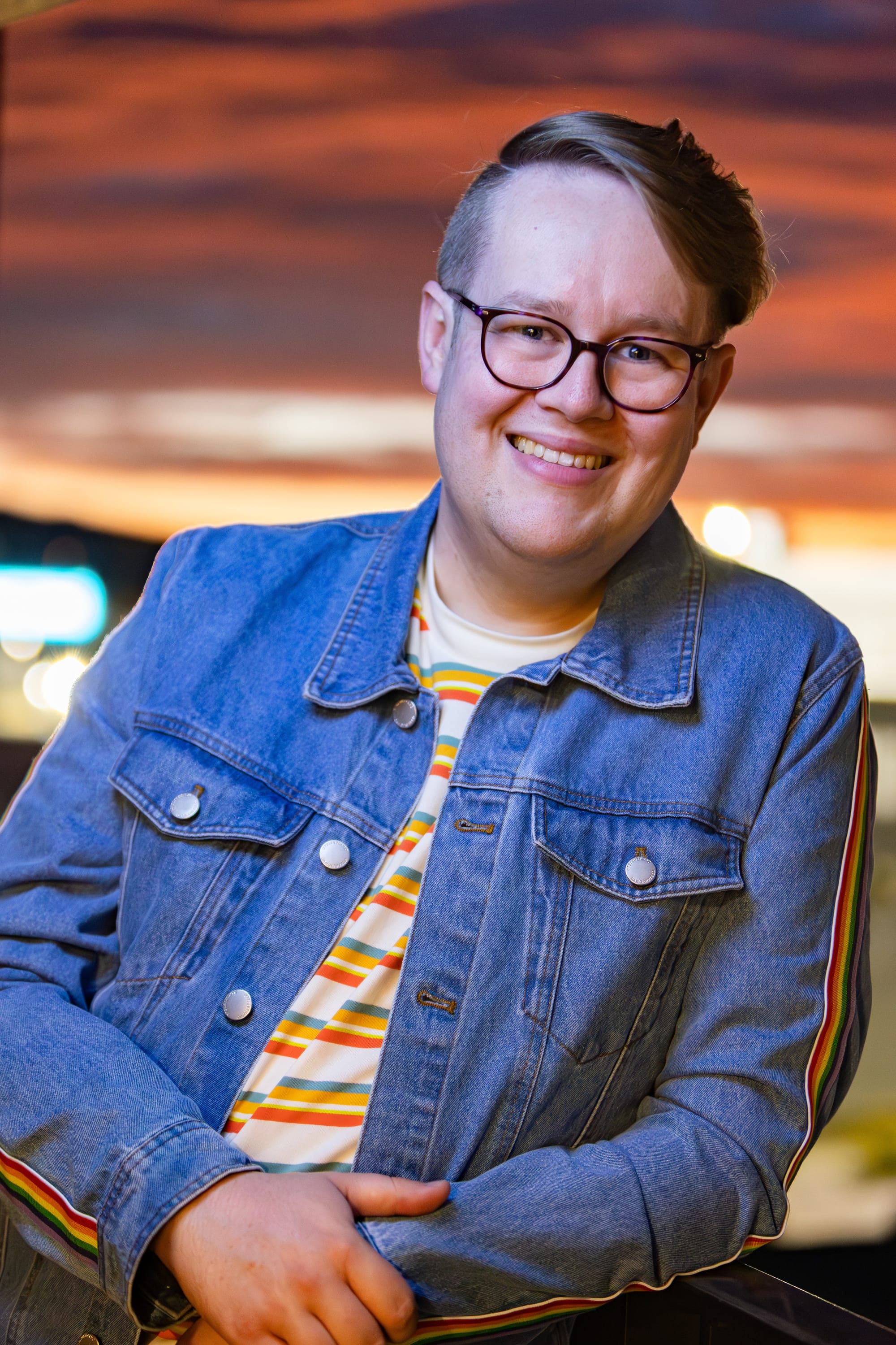 Mark smiling at the camera with a sunset and ember clouds in the background. He is smiling and wearing pink tortoise-shell glasses, a denim jacket with rainbow sleeve stripes, and a t-shirt with colourful stripes.